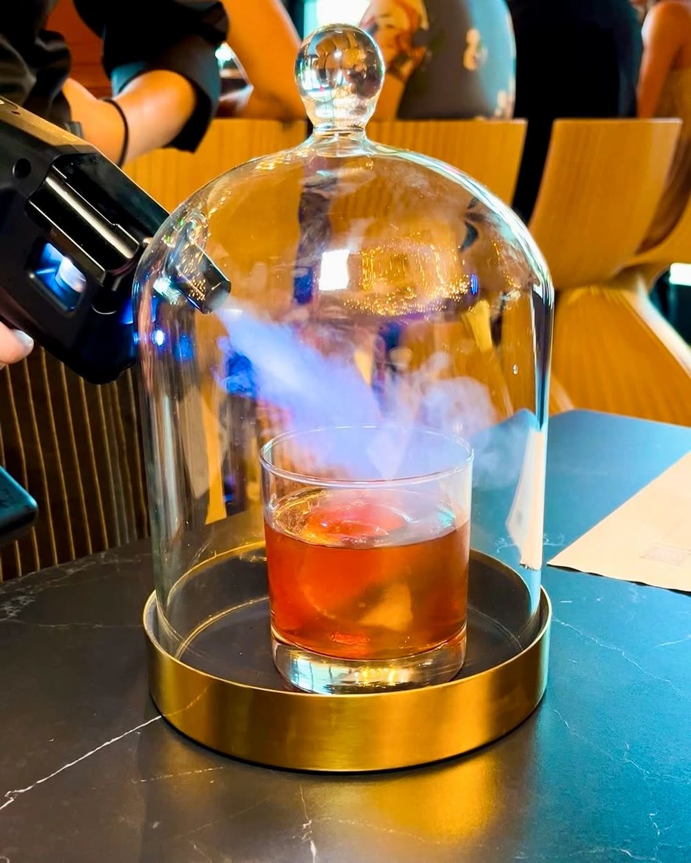 The Smoked Epic Fashioned being smoked under a glass cloche on a brass tray at the Tallo bar